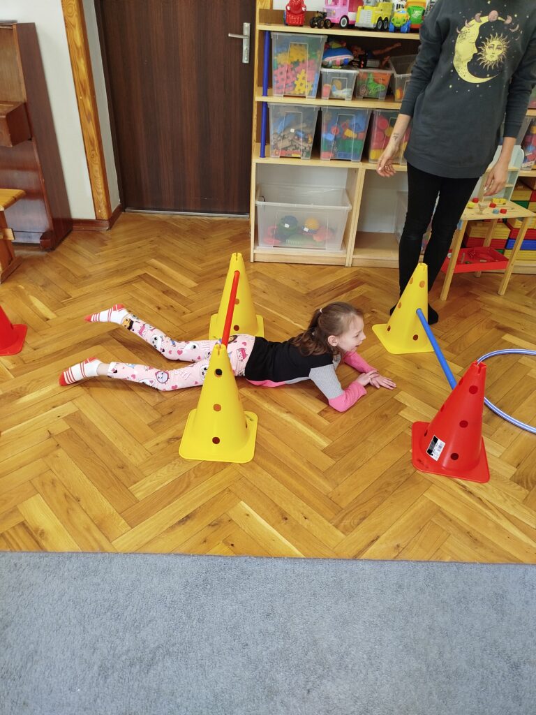 Dziewczynka w sali przedszkolnej czołga się po podłodze pod rozstawionymi palikami.