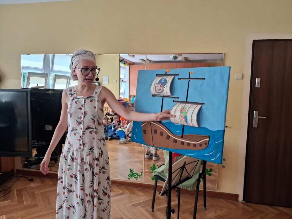Pani pokazuje rysunek papugi, który zamieszczony jest na plakacie ilustrującym satry statek.