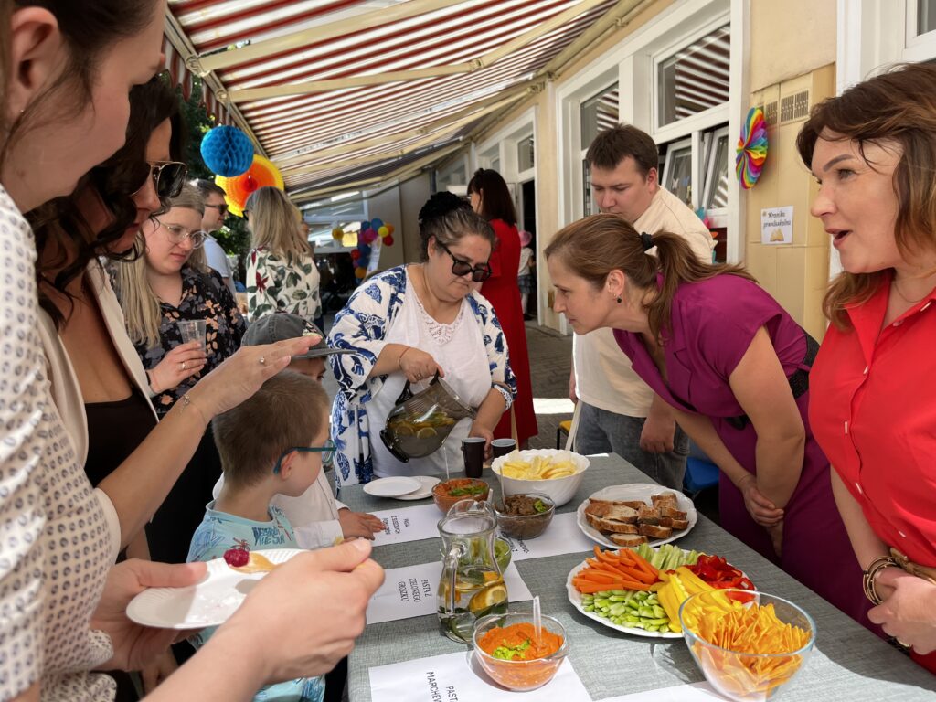 Stół z przysmakami przygotowanymi przez zespół kulinarny "Wiem, co jem" . Wokół stołu zebrani są nauczyciele i rodzice, którzy częstują się jedzeniem i piciem.