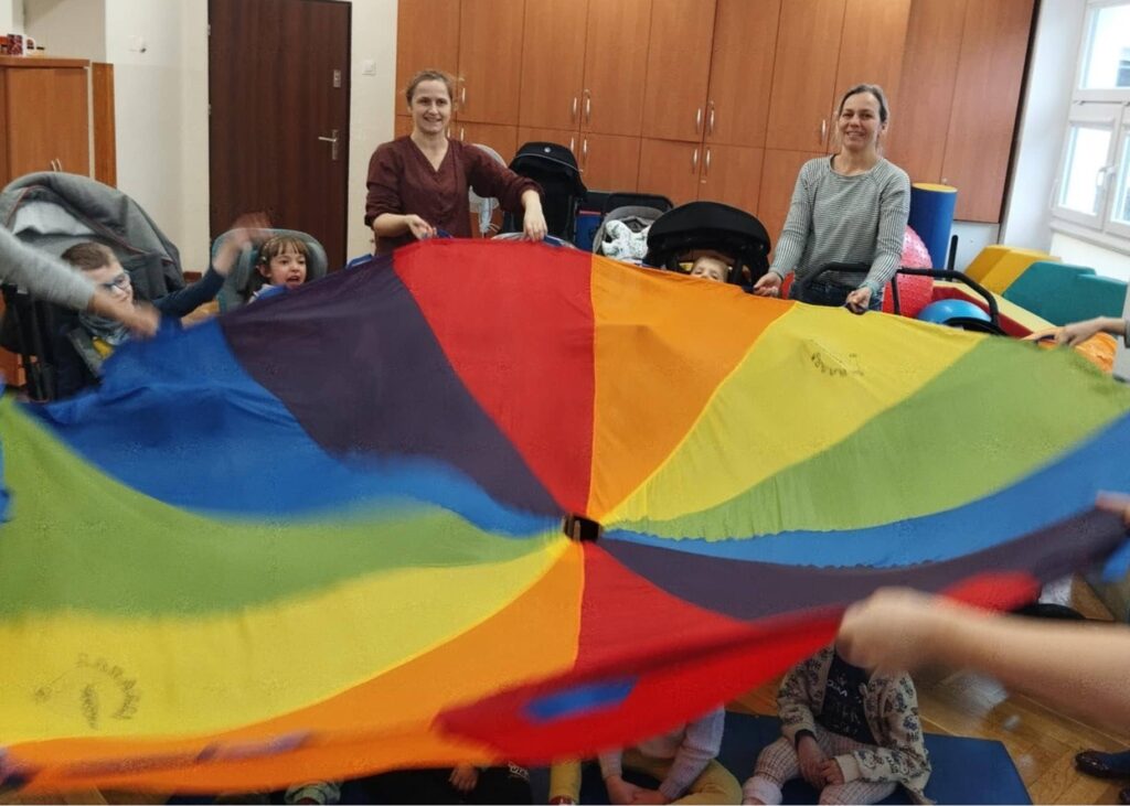 Dzieci i dorośli stoją w kole i trzymają kolorową chustę.