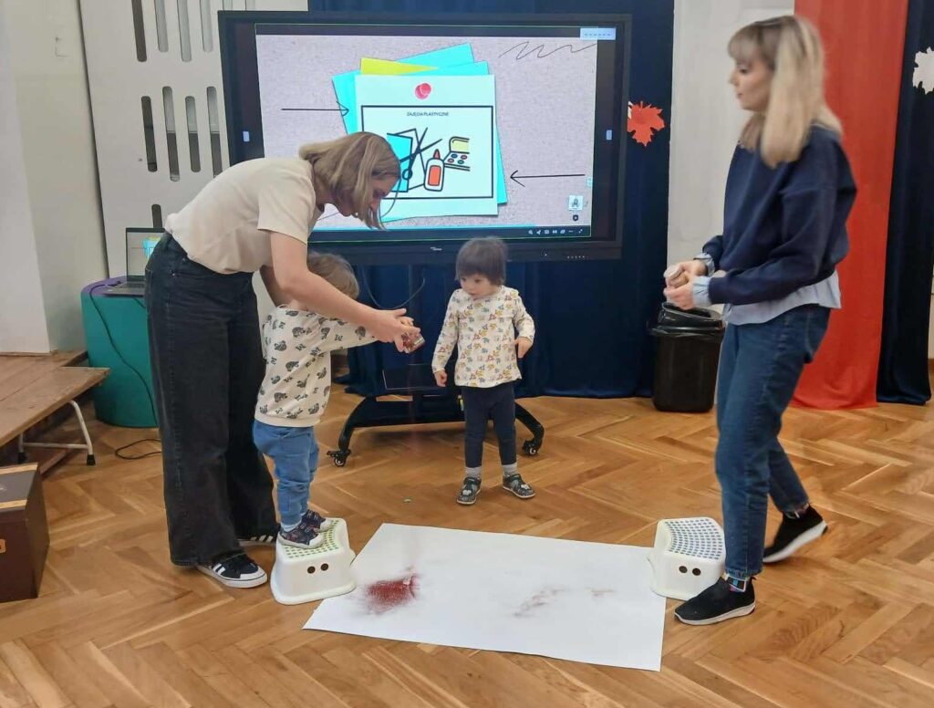 Dziecko stoi na podstawce i sypie piasek na kartkę papieru. Nauczyciel stoi z tyłu, za dzieckiem i trzyma jego rękę. W tle monitor z wyświetlanym obrazkiem PCS.