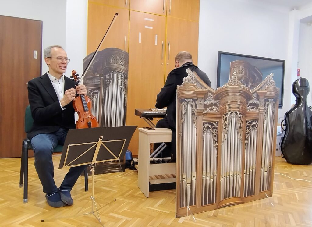 Muzyk trzyma skrzypce, obok stoi dekoracja organów, z tyłu drugi muzyk gra na keyboardzie.