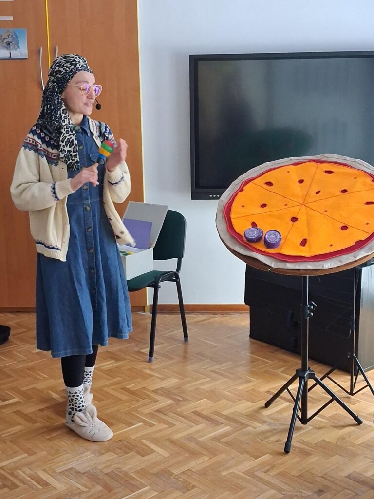 Pani stoi na środku sali, obok niej znajduje się makieta pizzy.